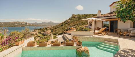 Una piscina con agua cristalina, rodeada de macetas con plantas y mobiliario exterior, con vistas a un paisaje pintoresco que incluye montañas y una masa de agua.