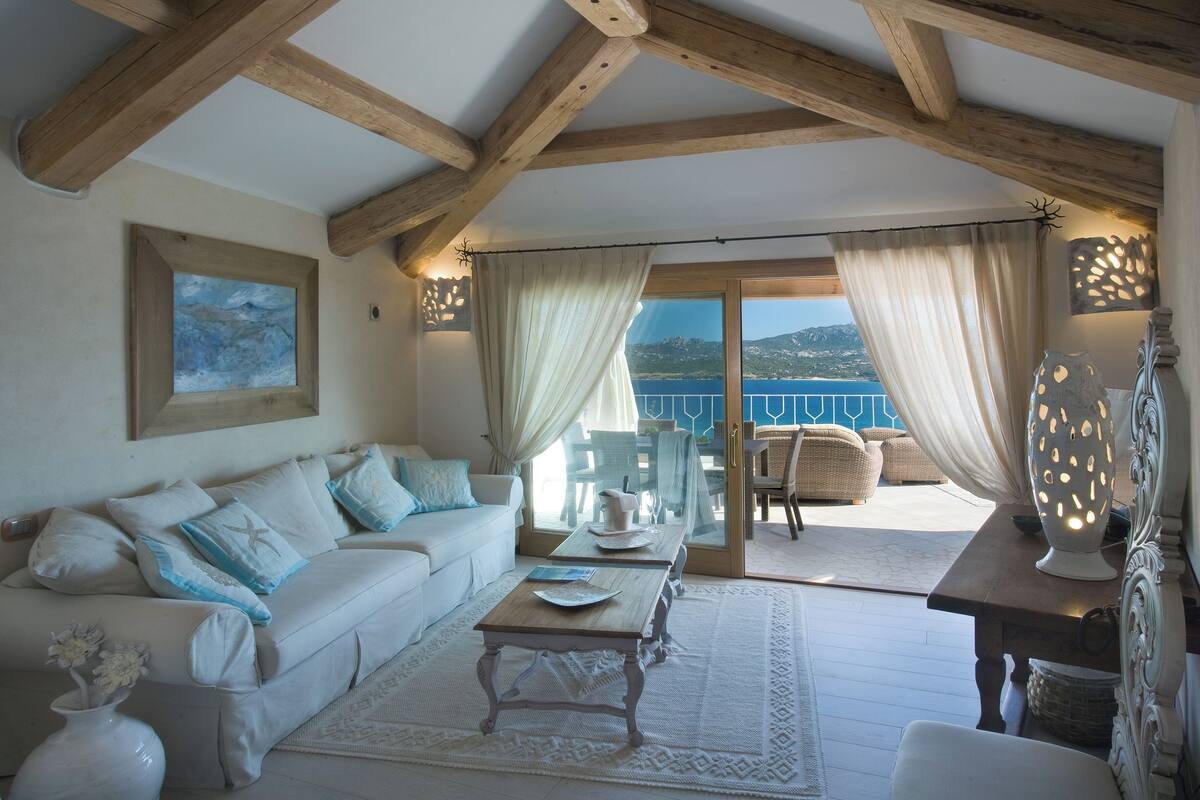 Ein gemütliches Wohnzimmer mit einem weißen Sofa, einem Holztisch und Meerblick durch große Glasfenster.