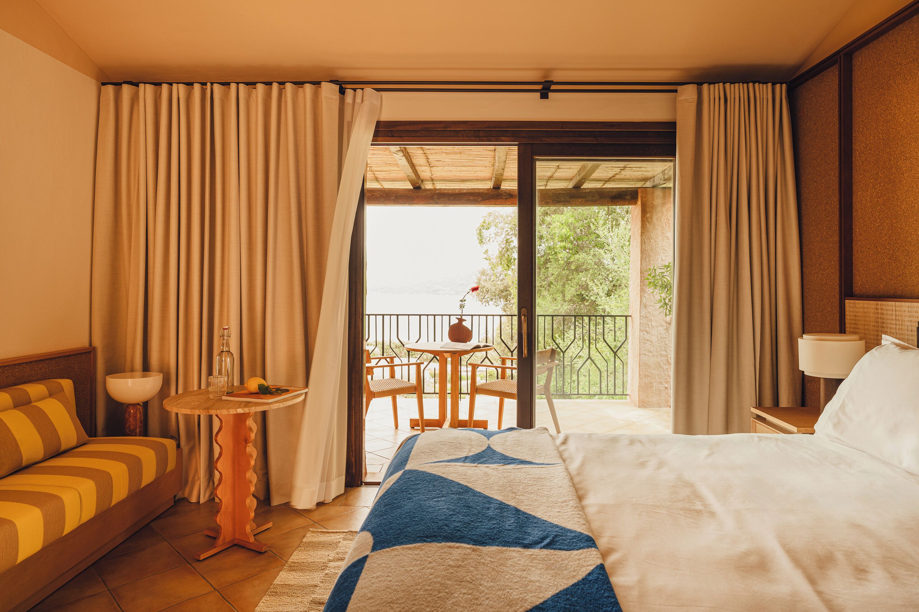 Een hotelkamer met een bed, een kleine tafel met een fles en glazen, een sofa en een balkon met een tafel en stoelen.