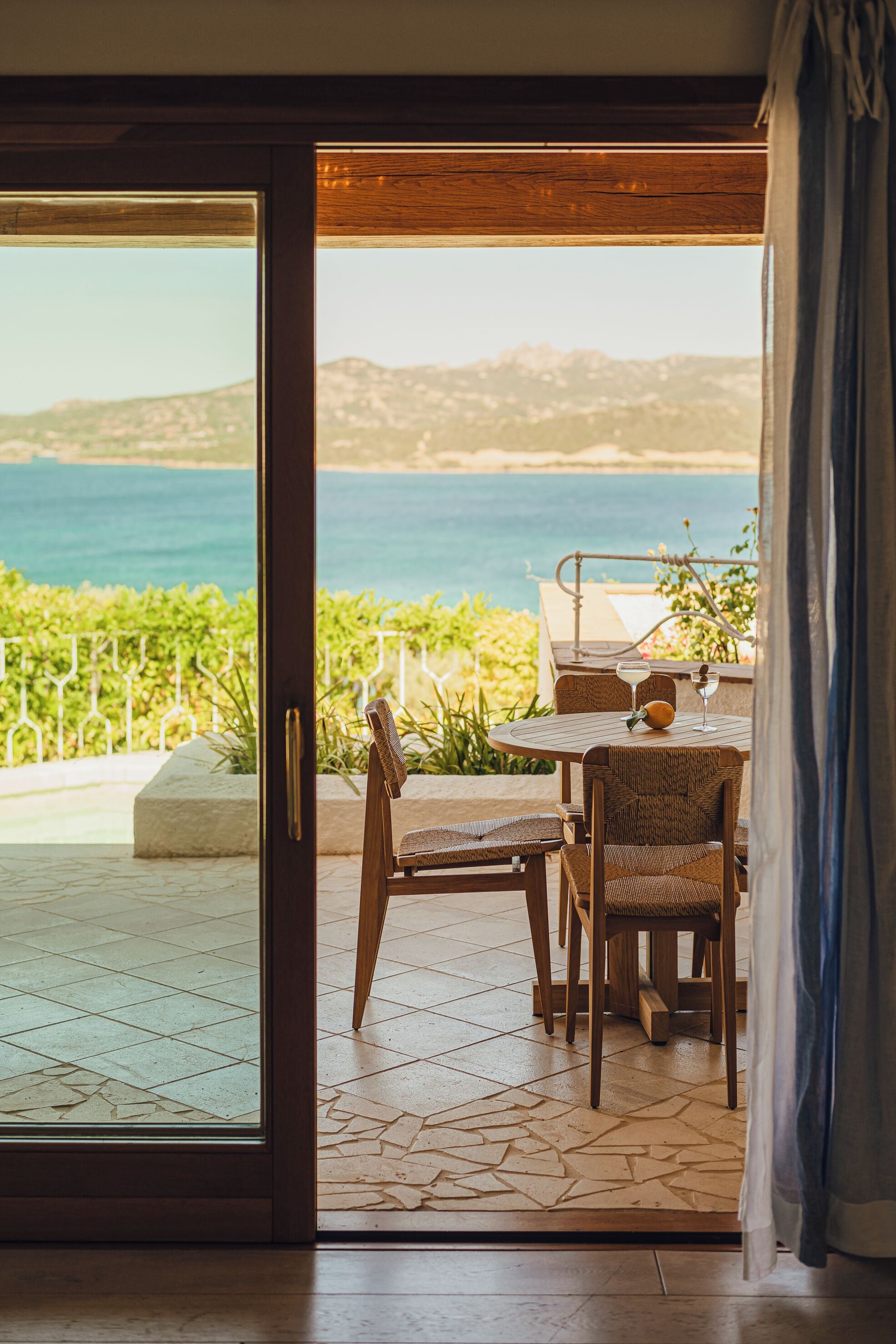 A room with a view of the sea, a table with chairs, and a glass of drink.