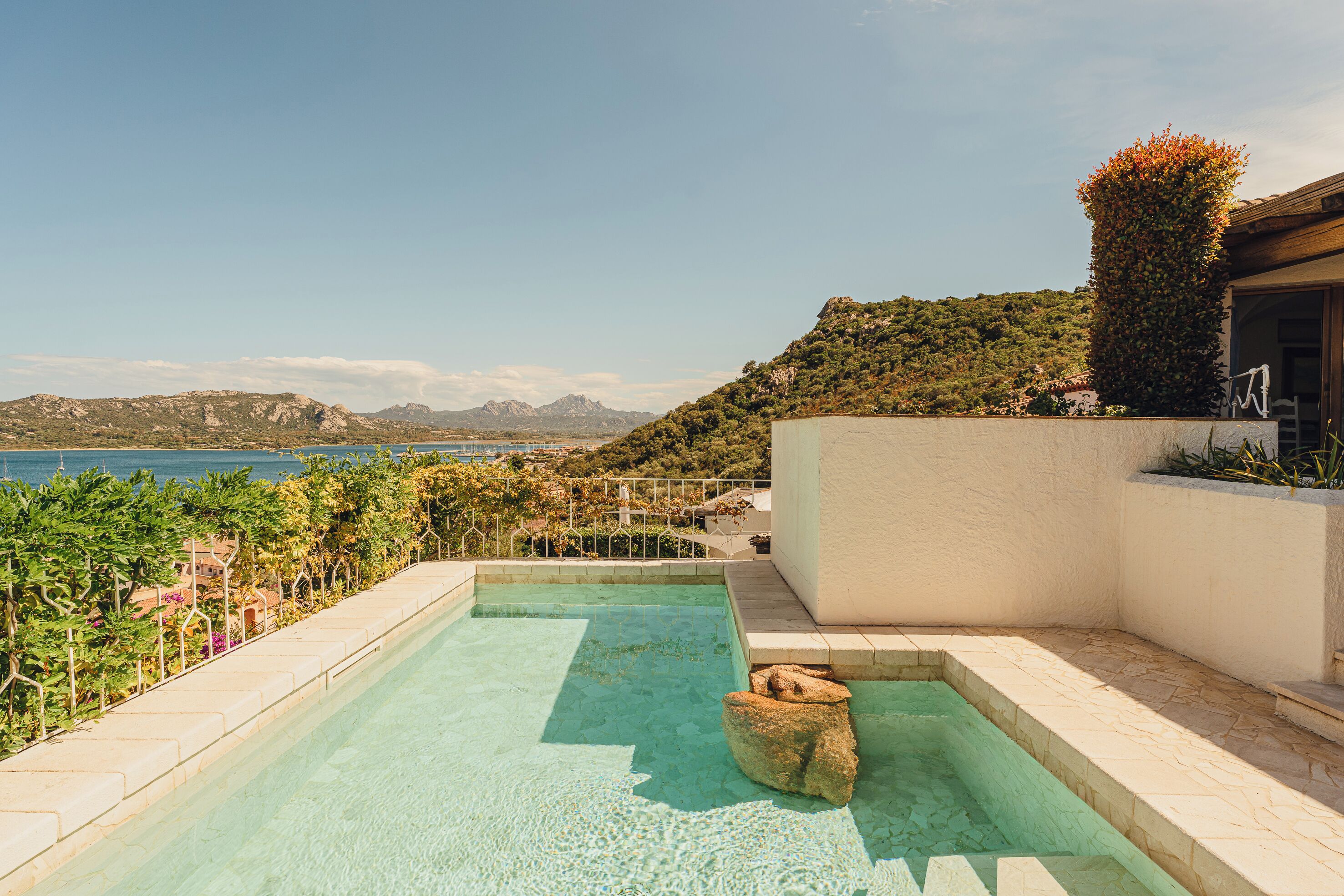Une piscine aux eaux cristallines, offrant une vue sur la mer et les montagnes, entourée d’une végétation luxuriante et bordée d’un bâtiment blanc.