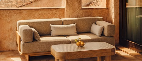 Un rincón exterior acogedor con un banco de madera y un sofá beige, una mesita con un frutero, y vistas a las montañas a través de un arco.