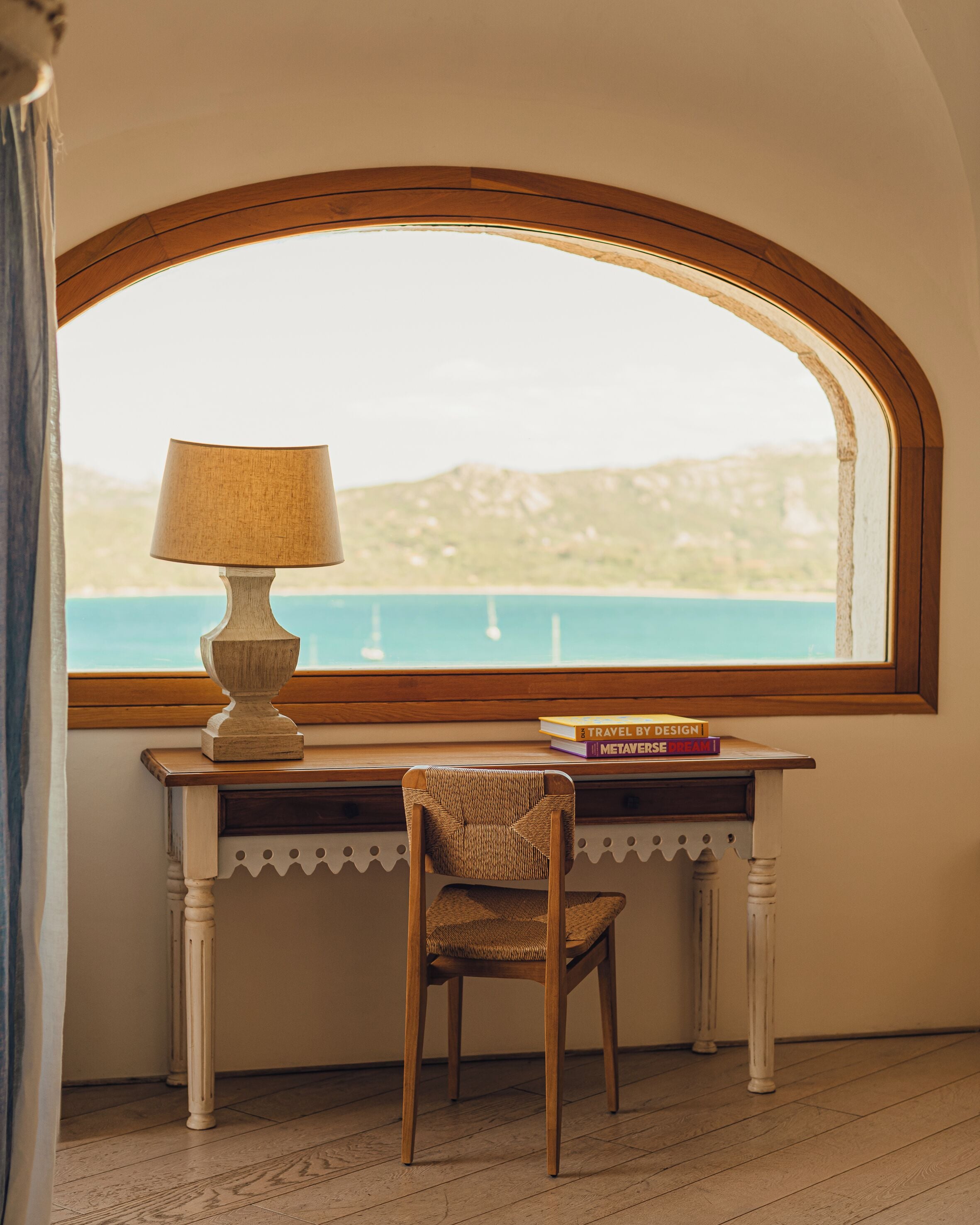 A desk with a lamp and books, a chair, and a view of the sea through an arched window.