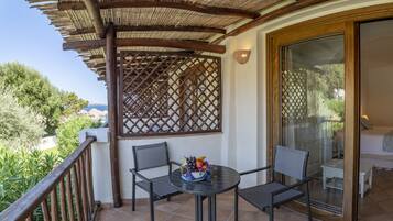 Ein Balkon mit Holzgitter, zwei Stühle, ein Tisch mit Obstschale und eine Glastür, die zu einem Schlafzimmer mit Bett und Sessel führt.