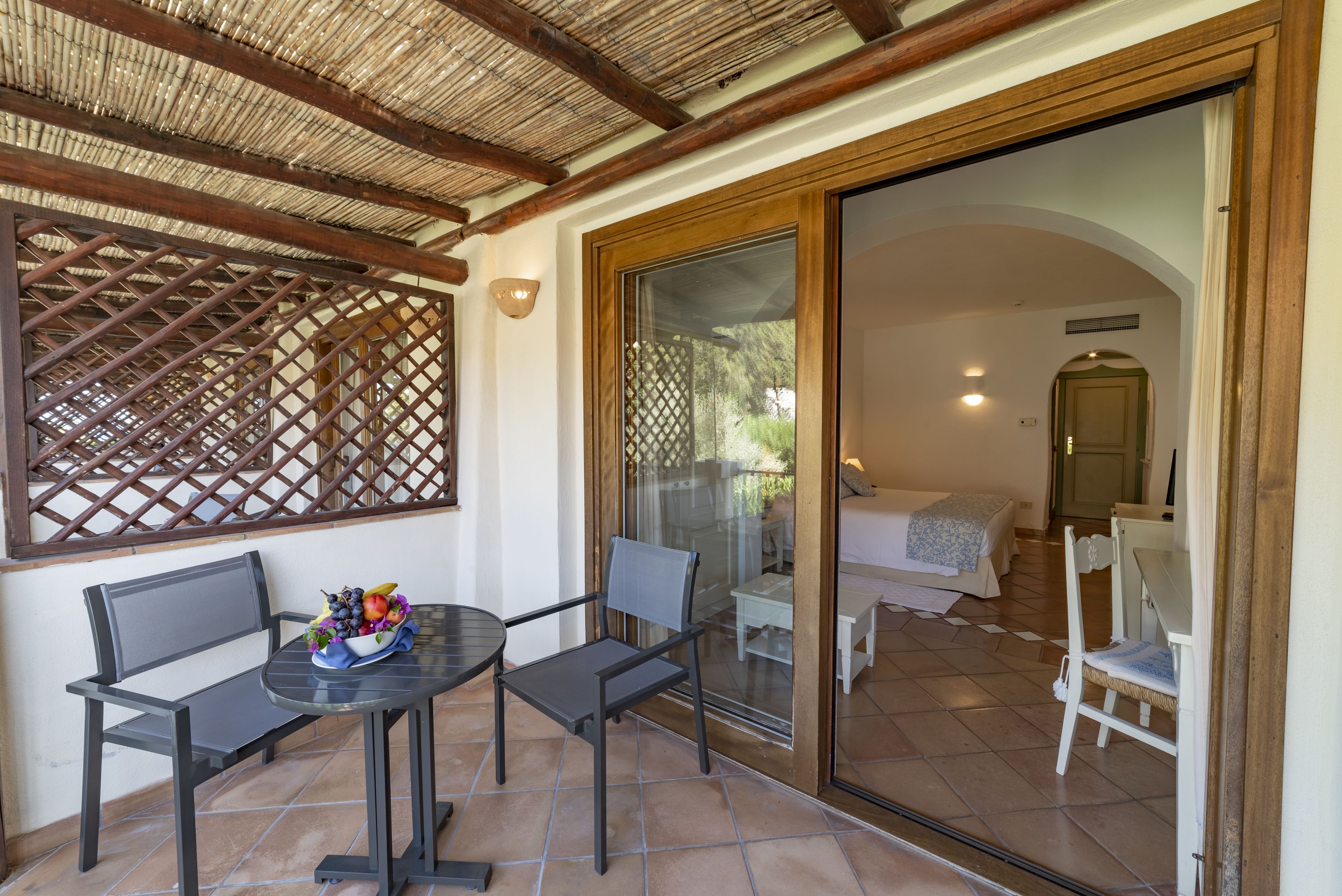 A patio area with a table and two chairs, a wooden lattice structure, and a sliding glass door leading to a bedroom with a bed and a nightstand.