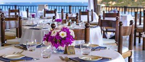 Una mesa lista para una comida con vista al mar, sillas de madera y un cartel con el número de la mesa.