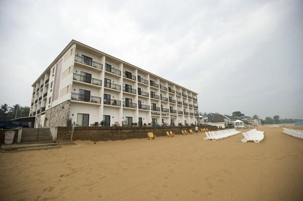 A multi-story hotel with balconies, a sandy beach in front, and beach chairs.