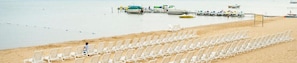 A beach with rows of white lounge chairs and a pier with boats in the background. - ParkShore Resort (Traverse City)