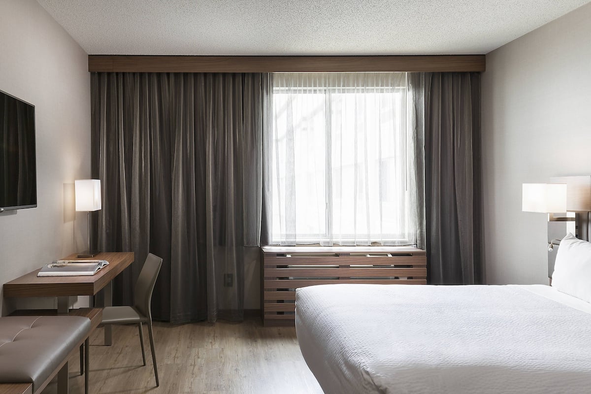 A hotel room with a bed, a desk, a chair, a window with curtains, and a TV on the wall.