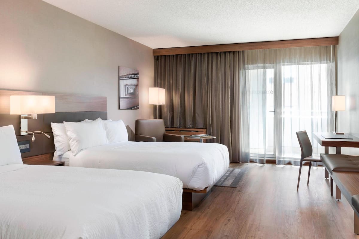 A hotel room with two beds, a desk, a chair, and a large window with curtains.