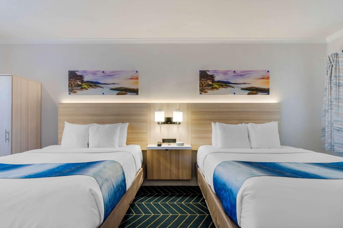 A hotel room with two beds, a nightstand with a lamp, and two wall-mounted paintings.
