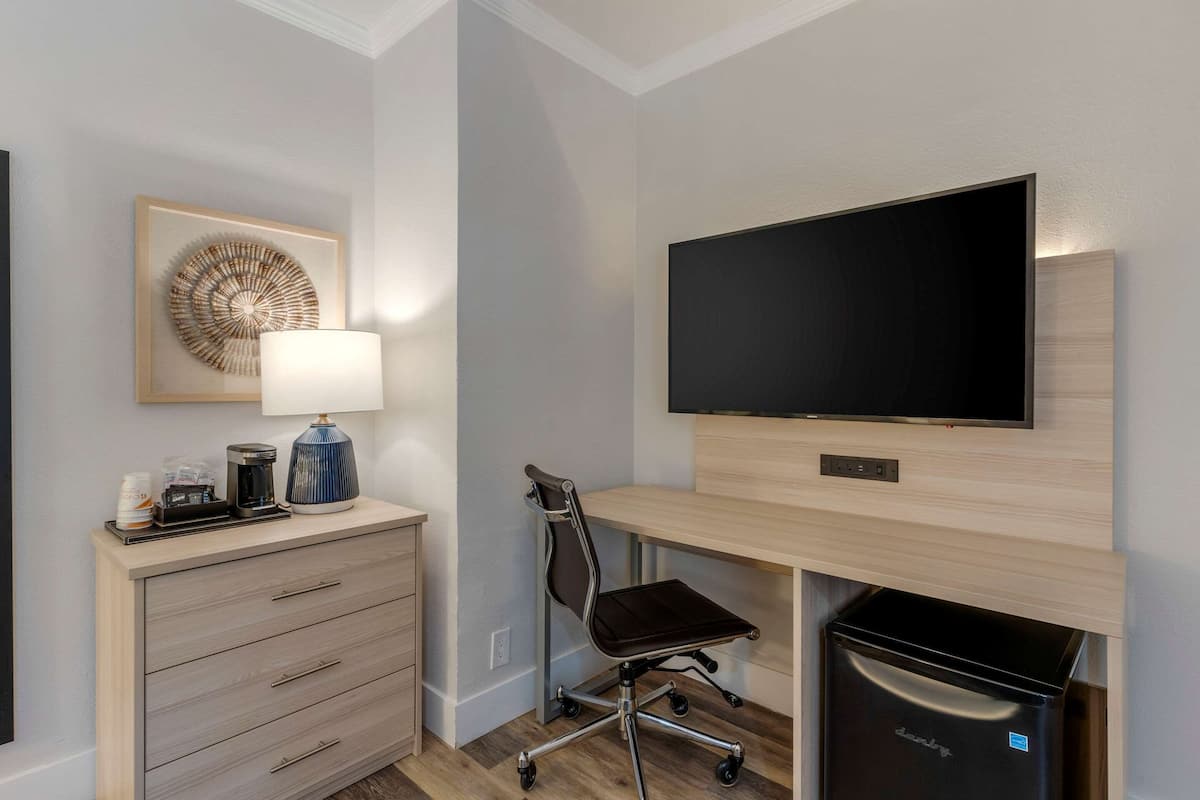 A modern hotel room with a wooden desk, a flat-screen TV, a lamp, a coffee maker, and a trash bin.