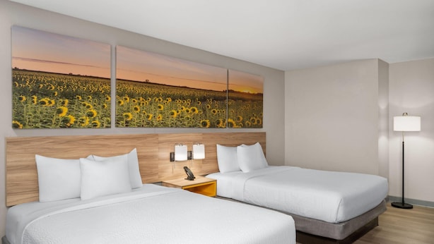 Habitación de hotel con dos camas, un cabecero de madera, una mesita de noche con lámpara y un cuadro grande de un campo de girasoles en la pared.