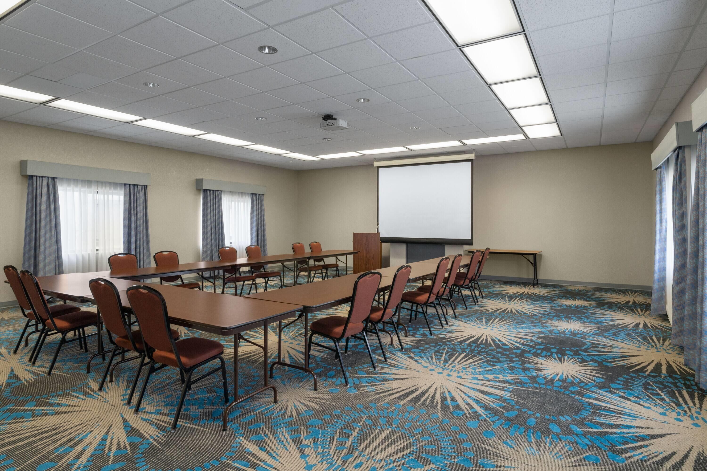 Un salón de conferencias con una larga mesa, sillas, una pantalla de proyector y una alfombra con diseño azul y blanco.