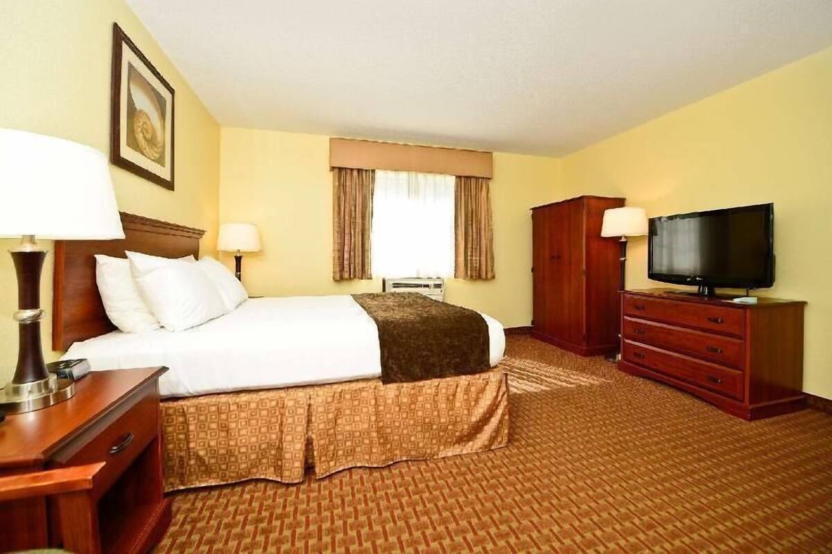 Quarto de hotel com cama, duas mesas de cabeceira, televisão, guarda-roupa e janela com cortinas.