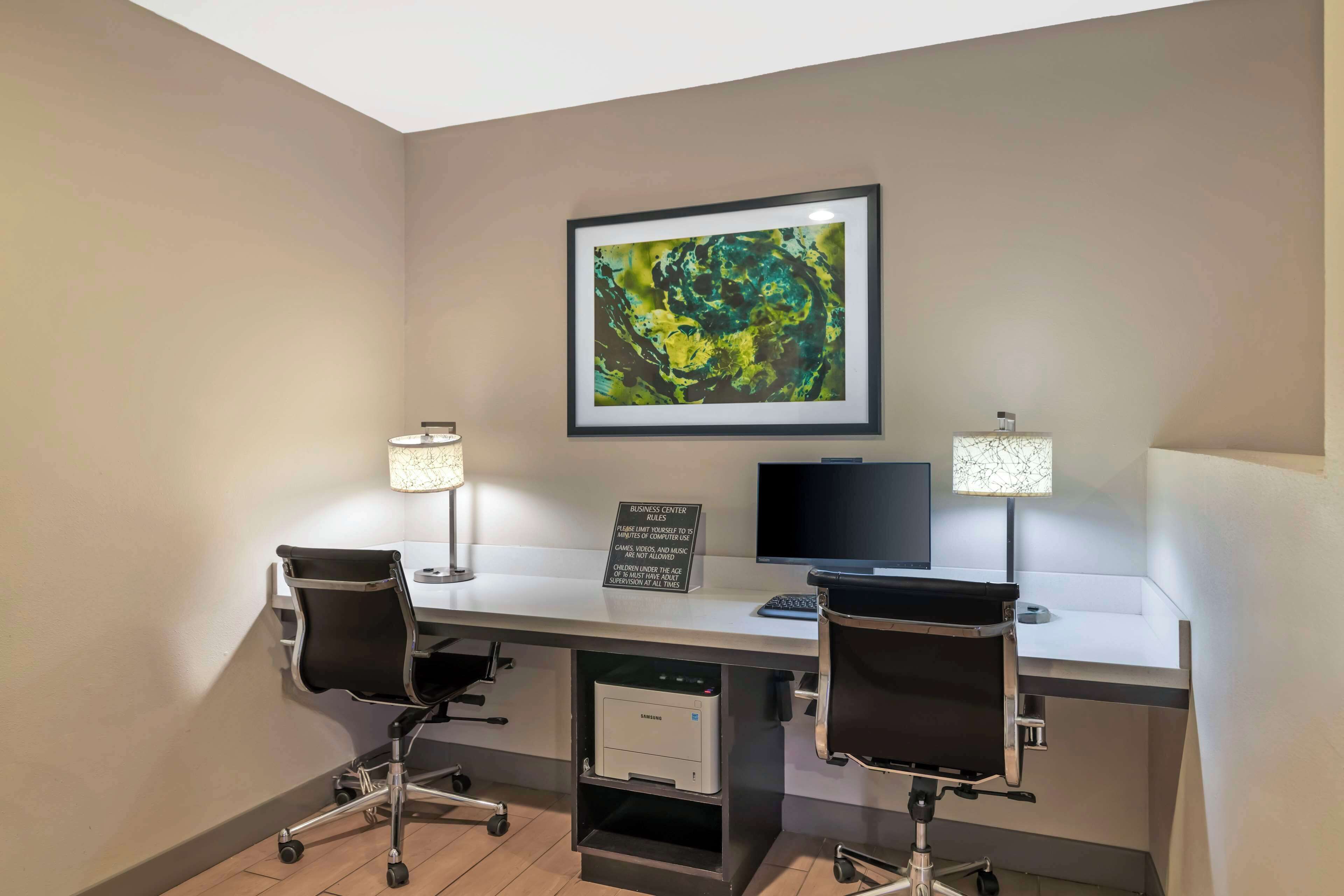 A business center with two desks, each with a chair, a monitor, and a lamp. There is a framed abstract painting on the wall and a sign indicating the business center's rules.