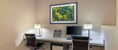 Un centro de negocios con dos escritorios, cada uno con una silla, un monitor y una lámpara. En la pared hay un cuadro abstracto enmarcado y un letrero que indica las normas del centro.