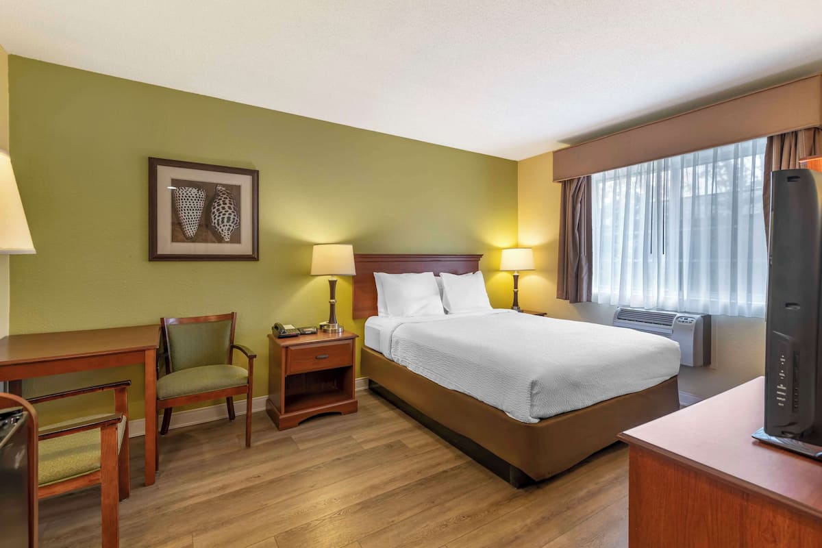 A hotel room with a bed, bedside lamps, a desk with a chair, a framed picture on the wall, and a window with curtains.