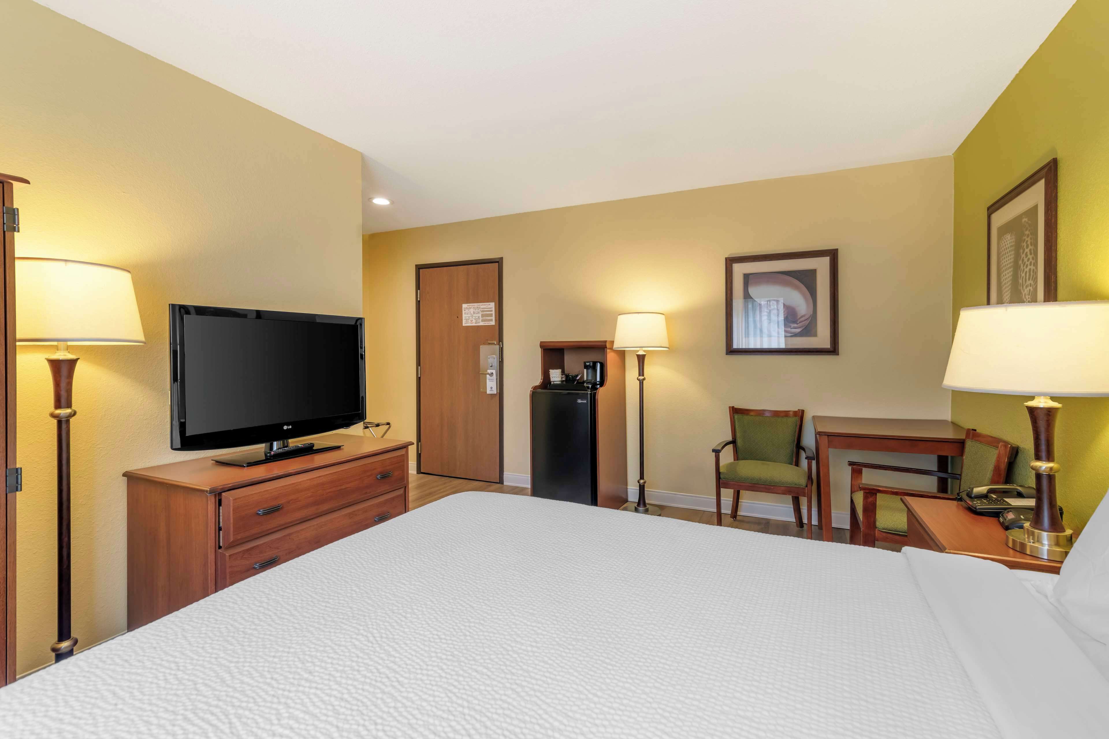 Habitación de hotel con una cama grande, un televisor en un aparador de madera, un escritorio con una silla, un refrigerador y dos lámparas.