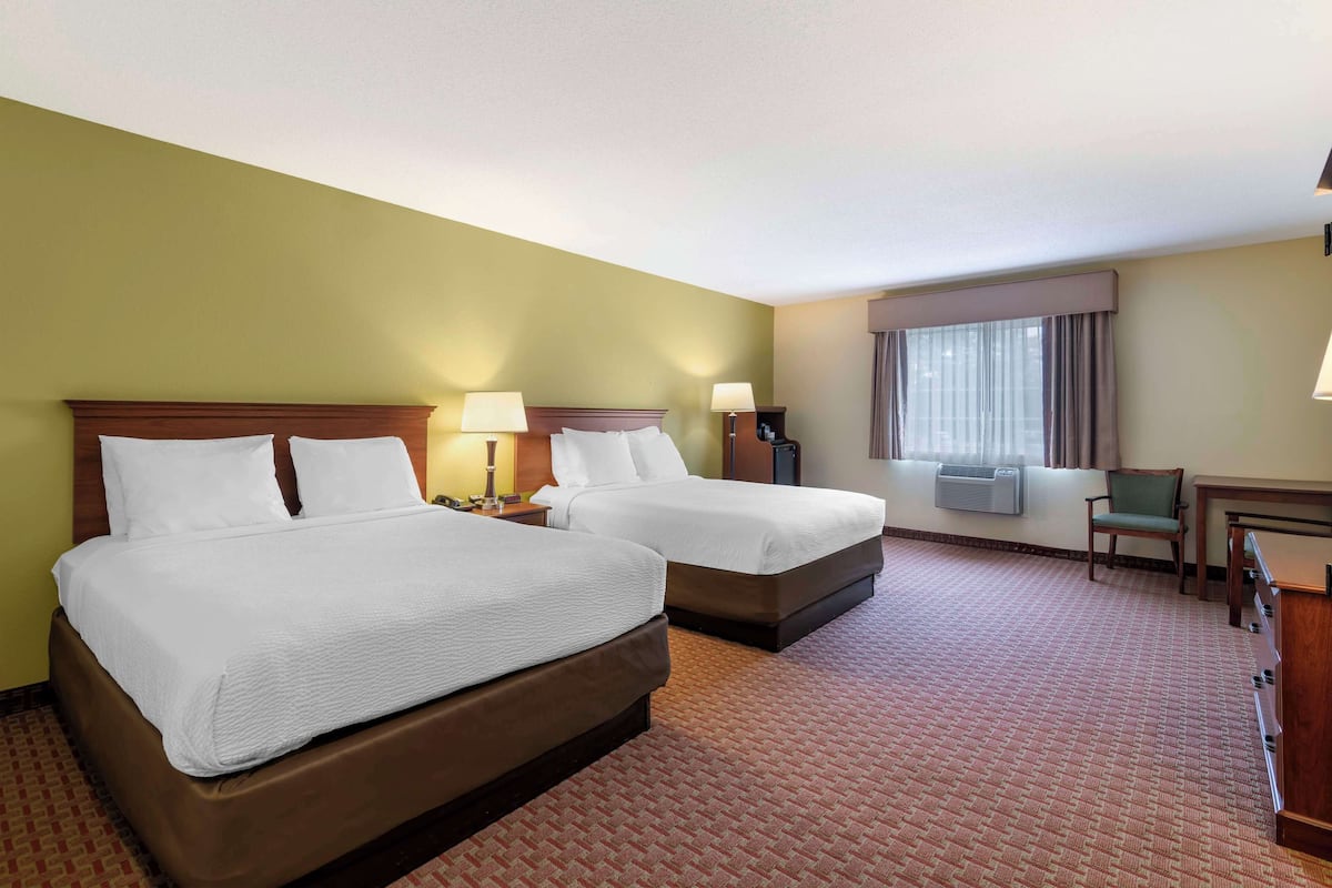 A hotel room with two beds, a desk, a chair, an air conditioning unit, and a window with curtains.