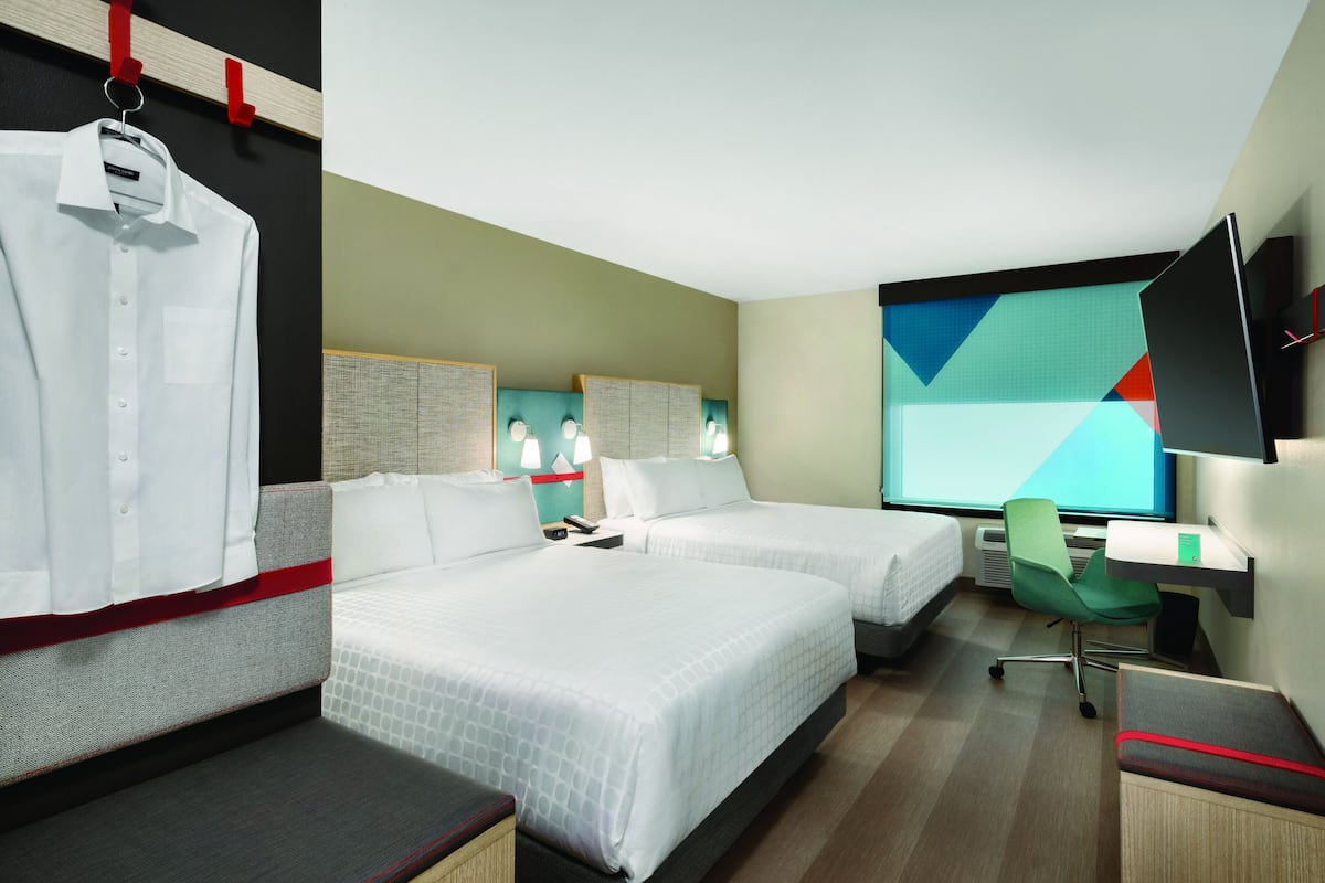 A hotel room with two beds, a desk, a chair, a TV, and a window with a geometric view.