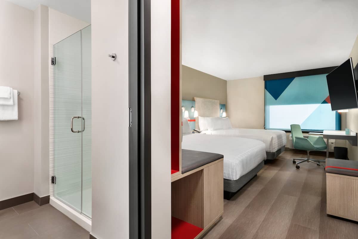 A modern hotel room with a glass shower enclosure, a bed with a red accent, a desk with a chair, and a large abstract painting on the wall.