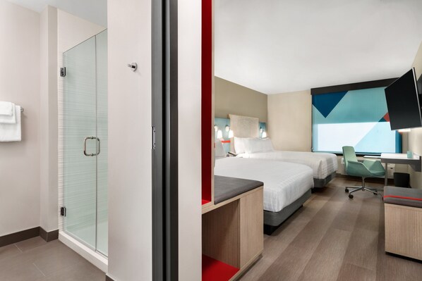 Una camera d'albergo moderna con cabina doccia in vetro, un letto con un tocco di colore rosso, una scrivania con una sedia e un grande quadro astratto appeso alla parete.