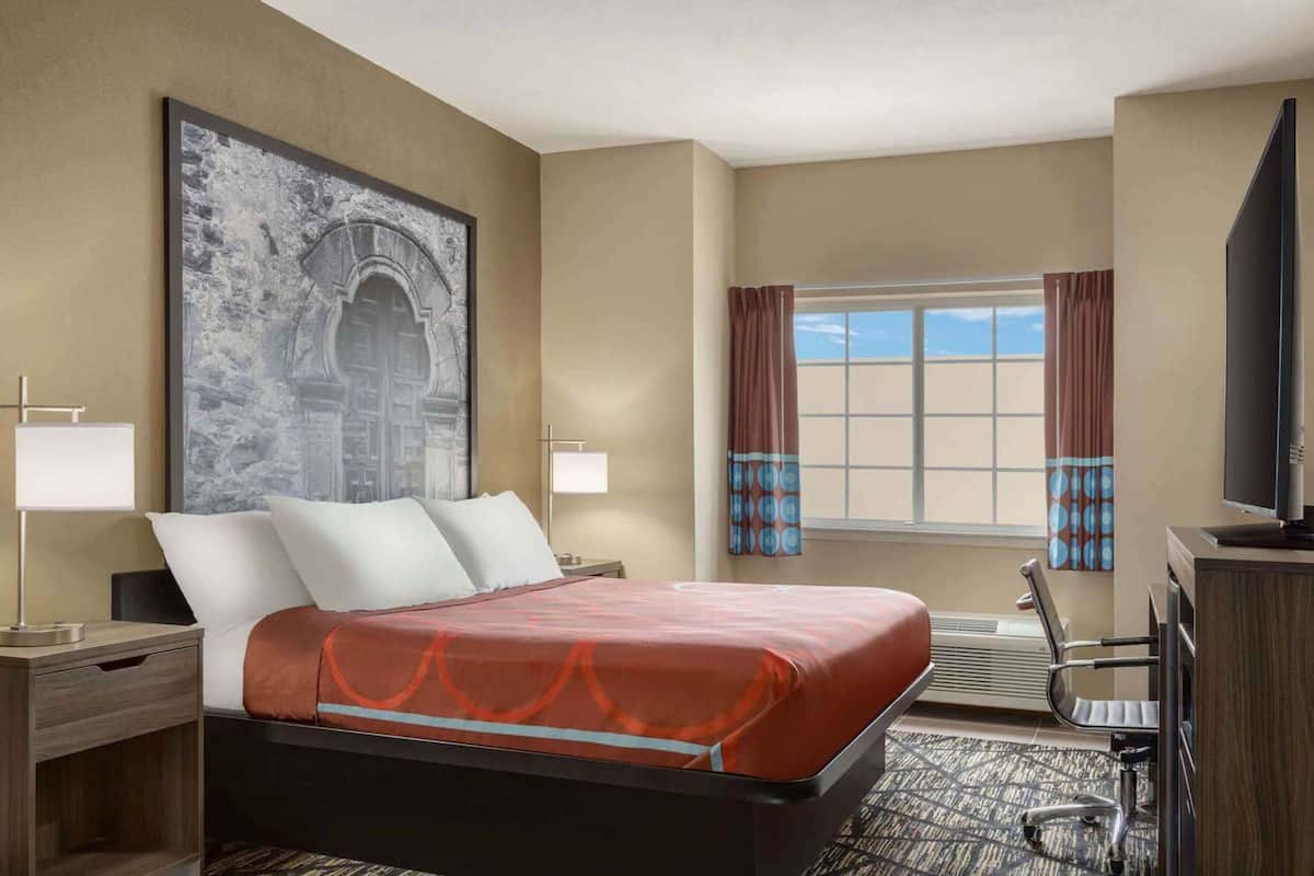 Une chambre d’hôtel avec un lit, un bureau, une chaise, une télévision, une fenêtre avec des rideaux et un tableau au mur.