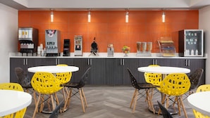 A modern kitchen area with a central island, equipped with a coffee machine, a microwave, and a refrigerator. There are four yellow chairs around the island, and the kitchen features a herringbone floor pattern.