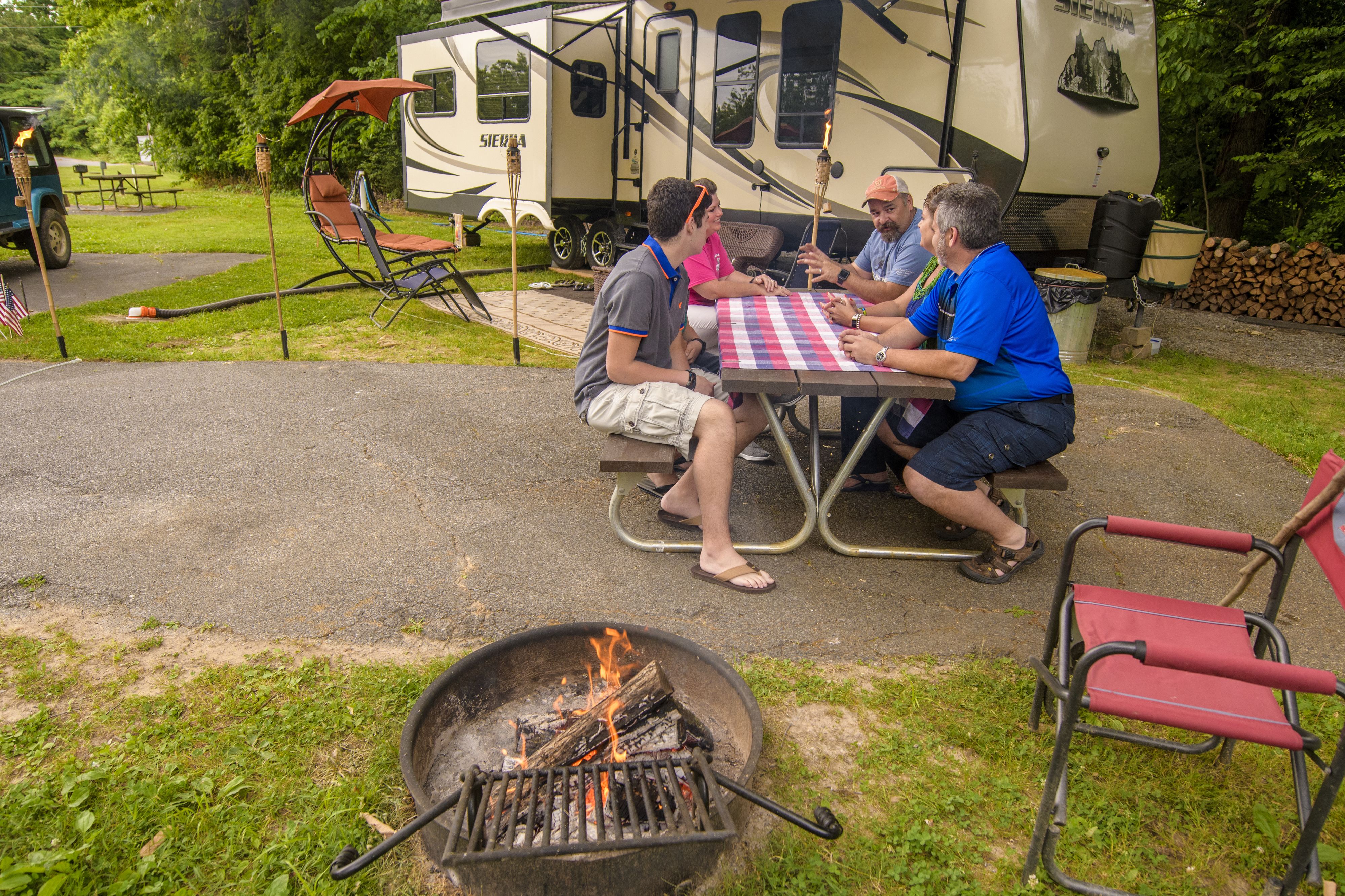 A group of people seated at a picnic table outdoors with a campfire nearby.