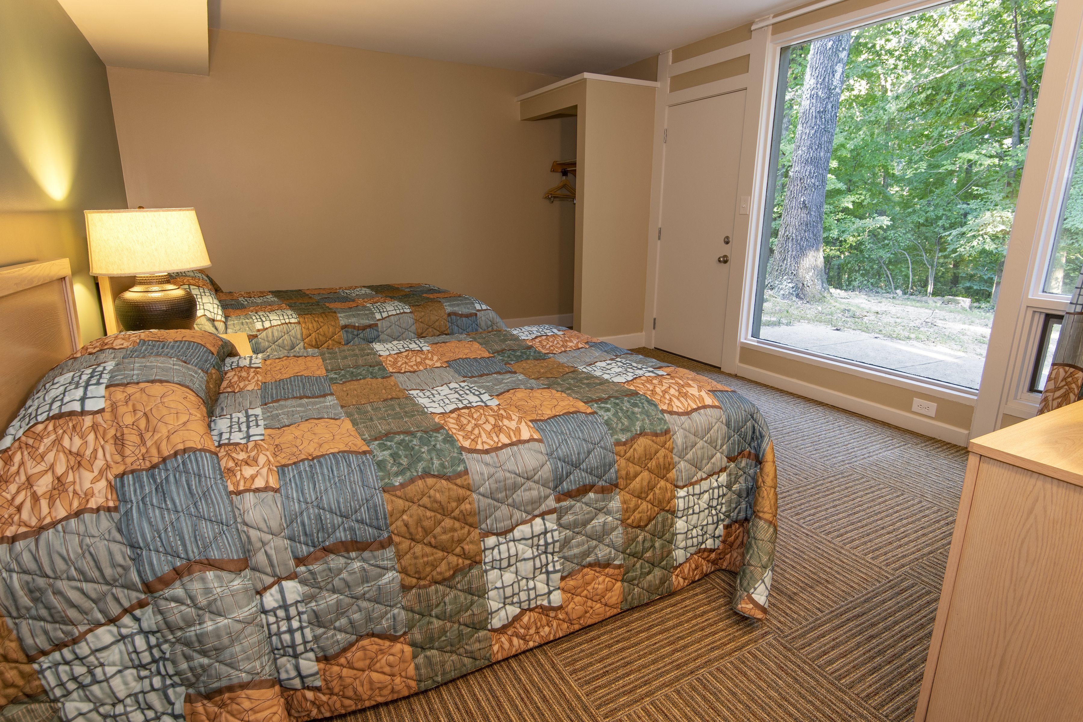 A hotel room with a large bed, a nightstand, a lamp, and a view of the outdoors through a sliding door.