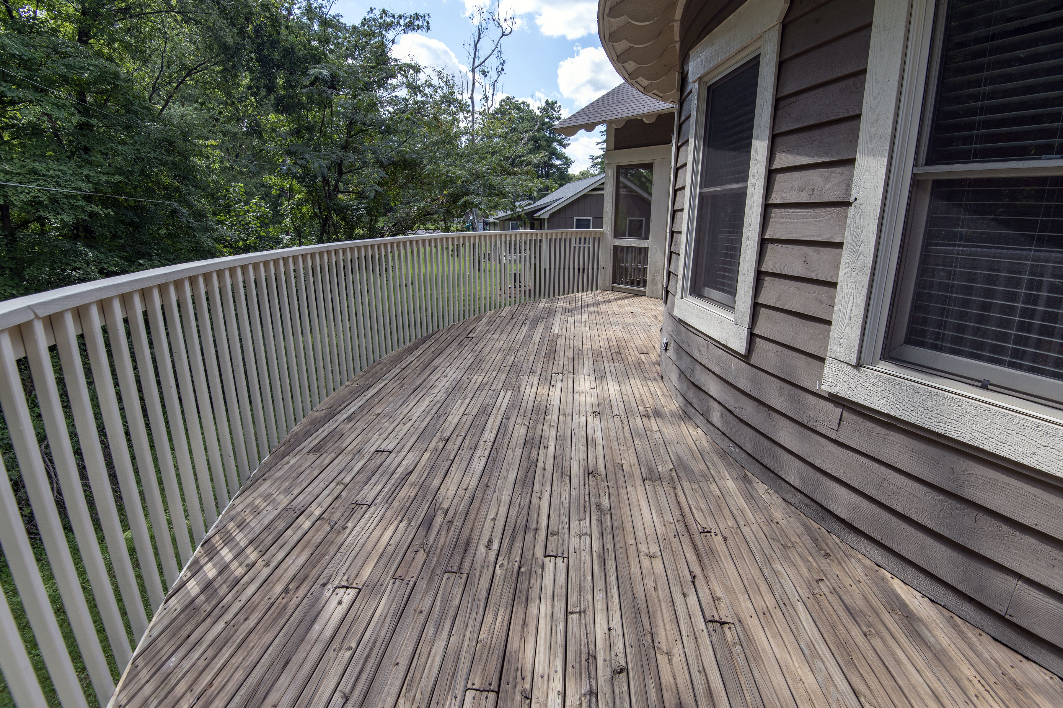 A wooden deck with a white railing, surrounded by trees and a house with large windows.