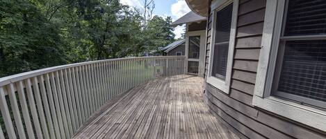 A wooden deck with a white railing, surrounded by trees and a house with large windows.