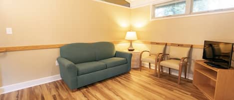 A room with a green sofa, wooden flooring, a lamp, two chairs, a TV on a wooden stand, and a window with blinds.