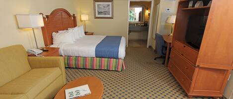 A hotel room with a bed, a sofa, a TV, a desk, and a bathroom visible through an open door.