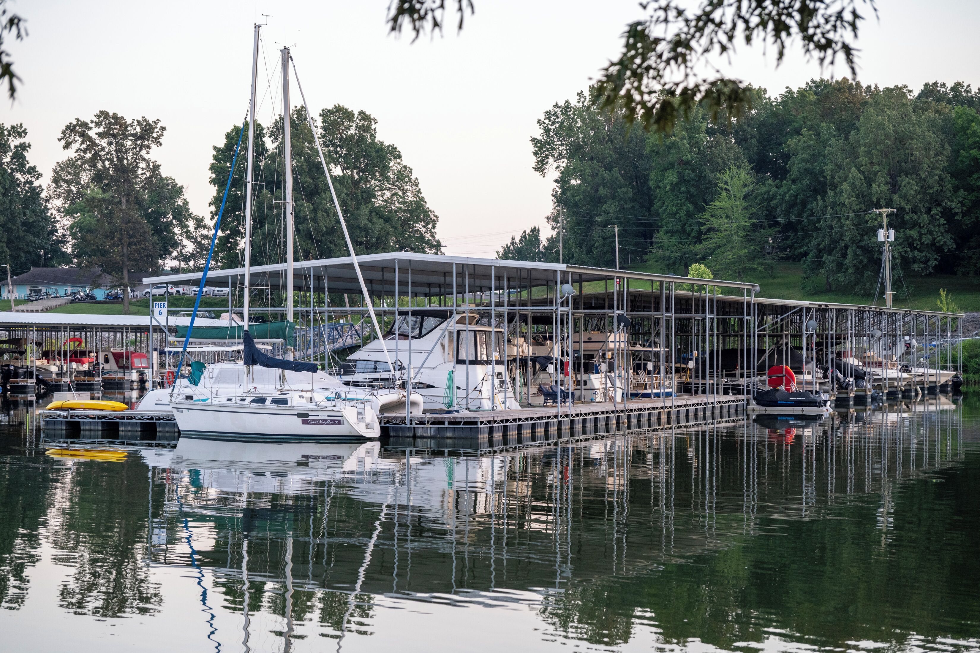 Een jachthaven met aangemeerde boten, een overdekte constructie en een gazon met bomen op de achtergrond.