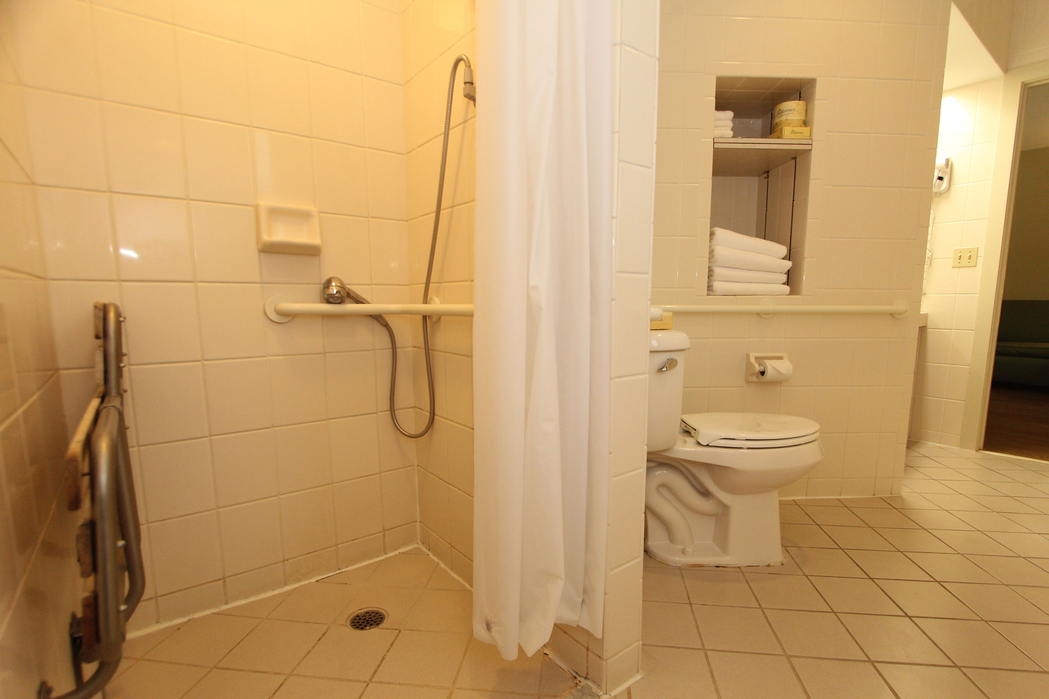 A bathroom with white tiles, a shower with a grab bar, a toilet, and a shelf with towels.