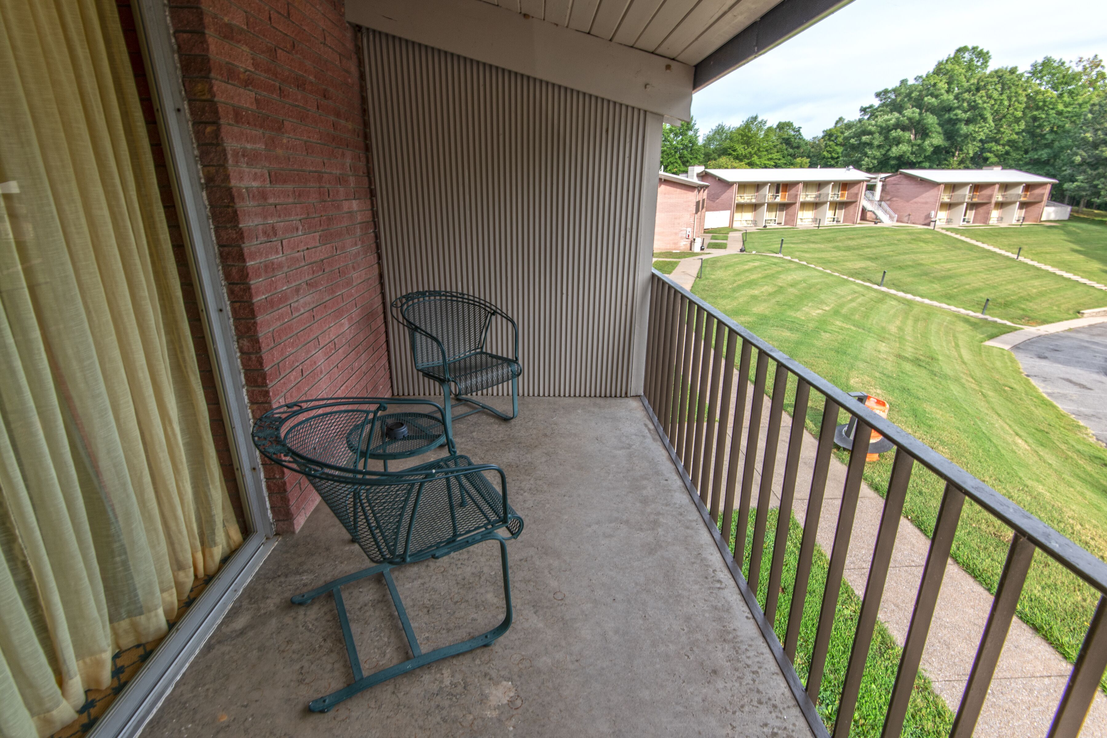 A balcony with two metal chairs, a brick wall, and a view of a green lawn and buildings in the background.