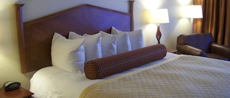 A hotel room with a large bed, multiple pillows, a bedside lamp, a chair, and a nightstand with a clock.