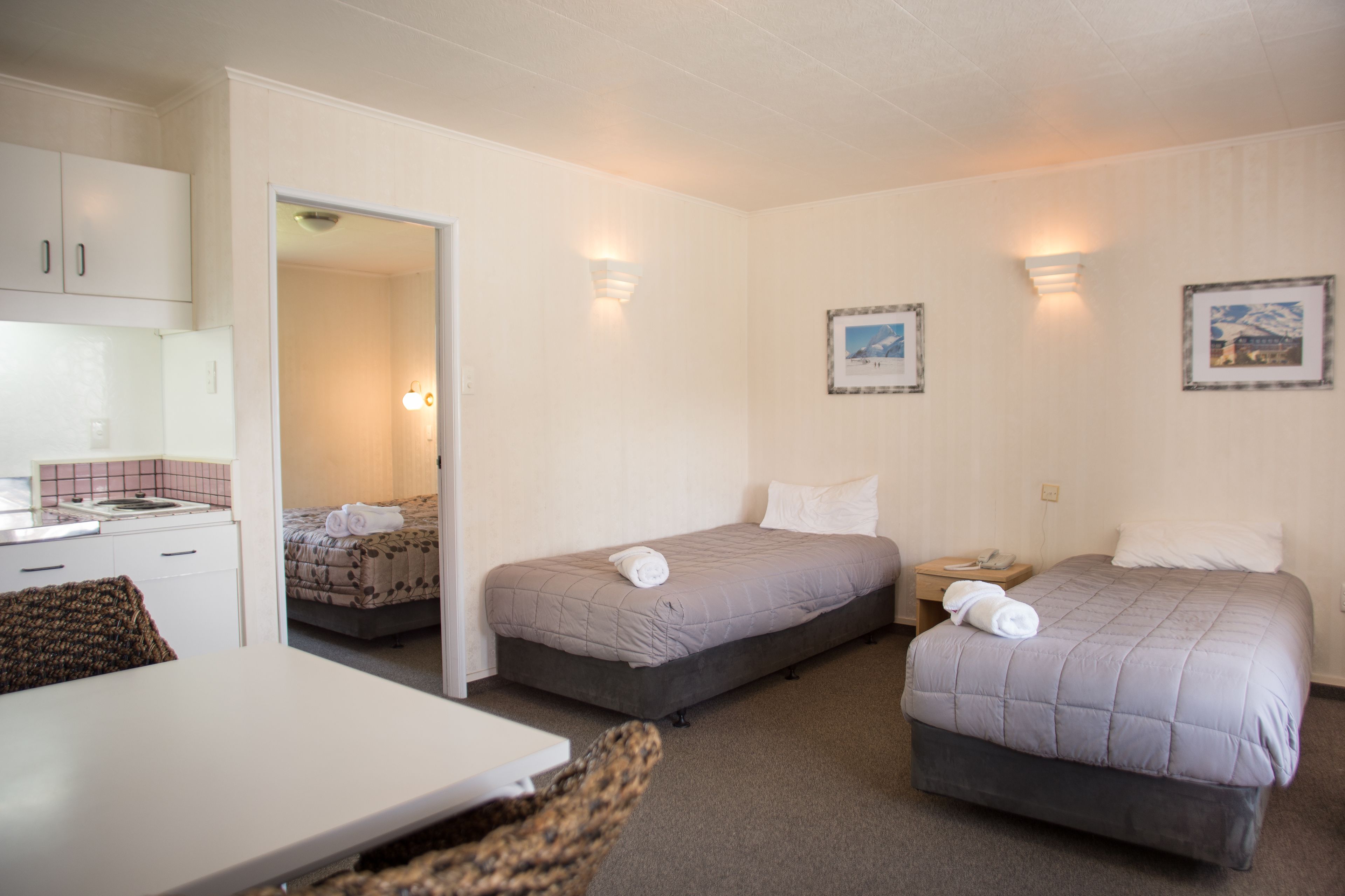 Habitación de hotel con dos camas, una mesita, una silla y una kitchenette.
