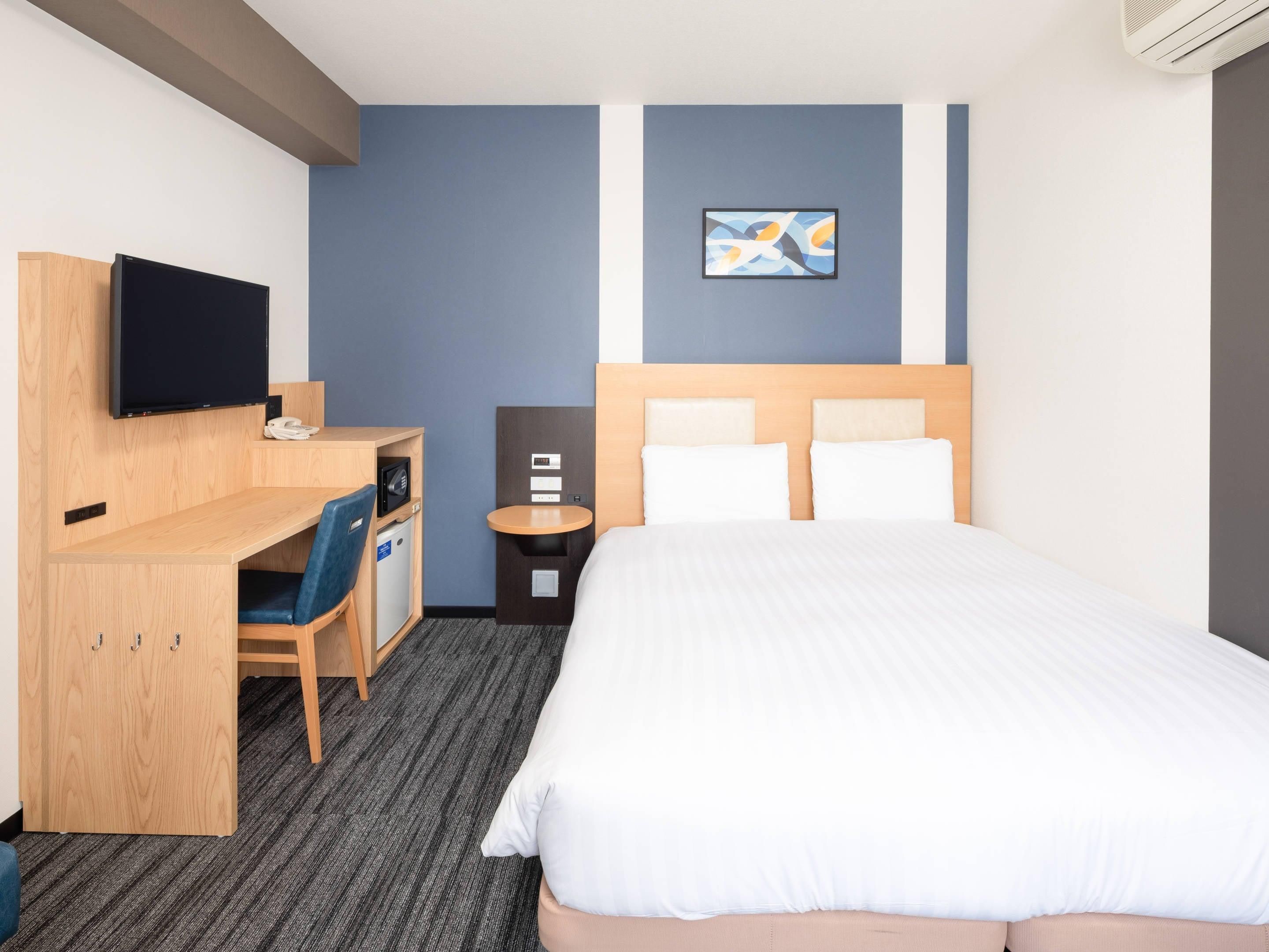 A hotel room with a bed, a desk with a chair, a television, a small table, and a wall-mounted artwork.