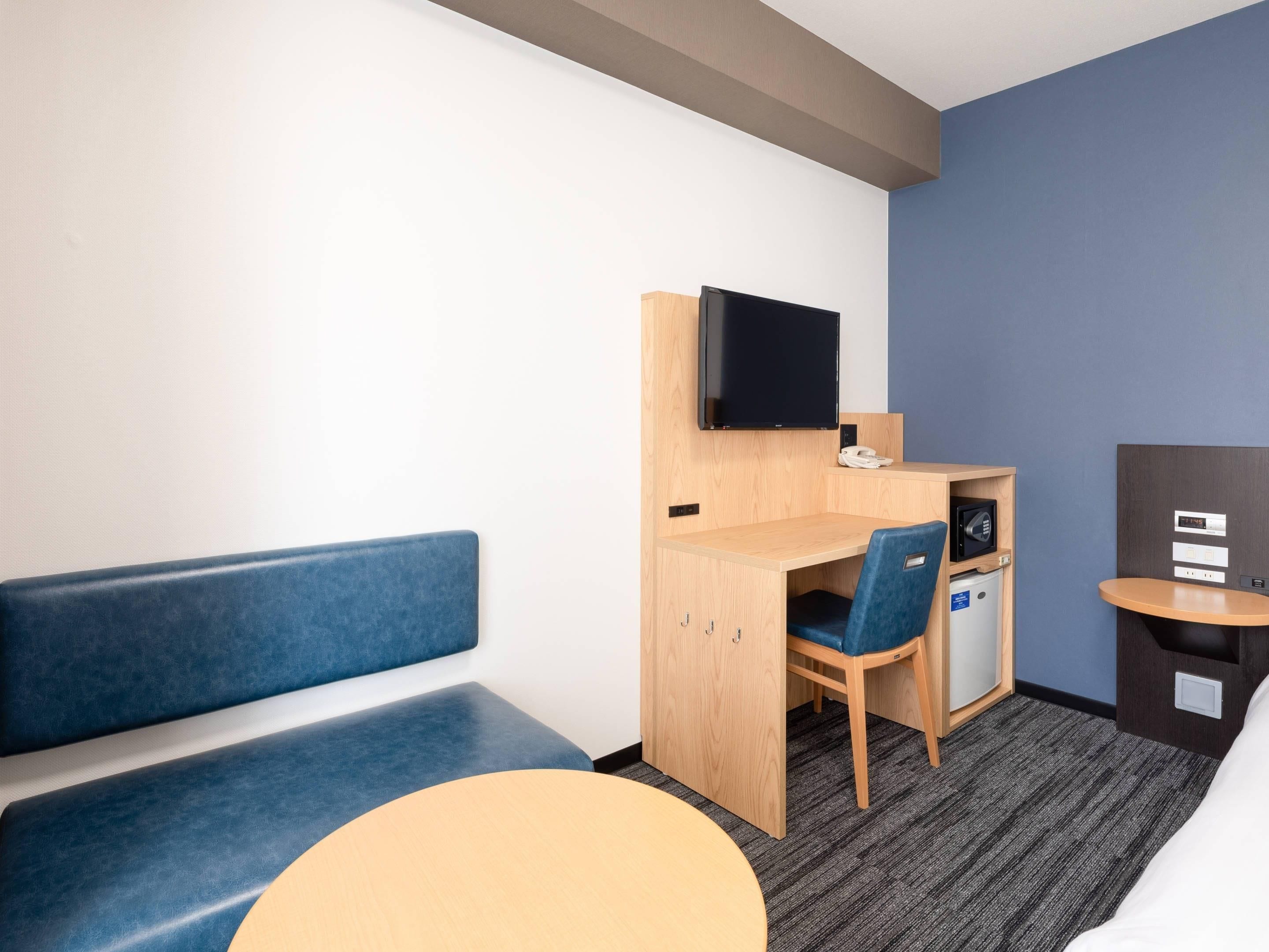 Habitación de hotel con cama, escritorio, silla, televisor, teléfono y una pequeña caja fuerte.