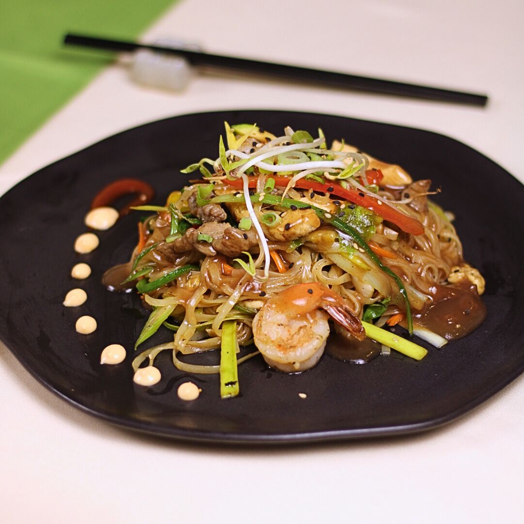 A plate of stir-fried noodles with vegetables and shrimp, garnished with sesame seeds and served on a black plate.