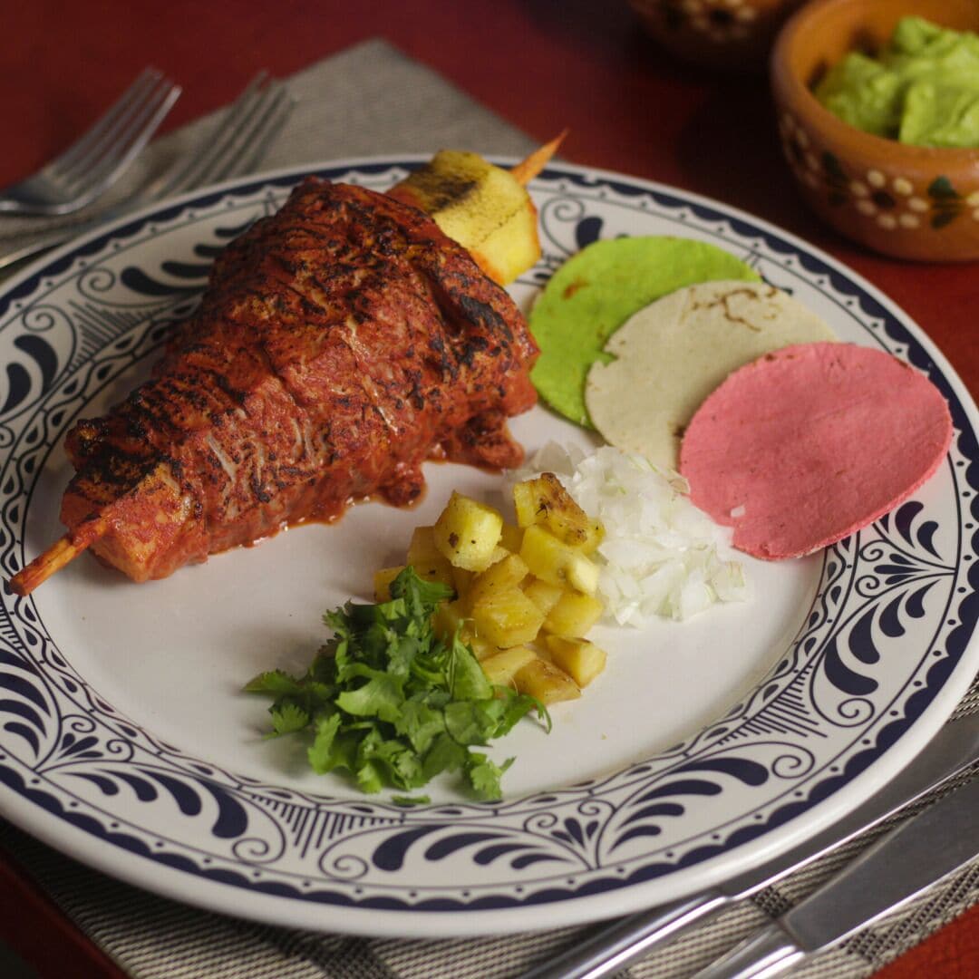 A plate with a grilled meat skewer, rice, a slice of meat, and a side of pineapple chunks.