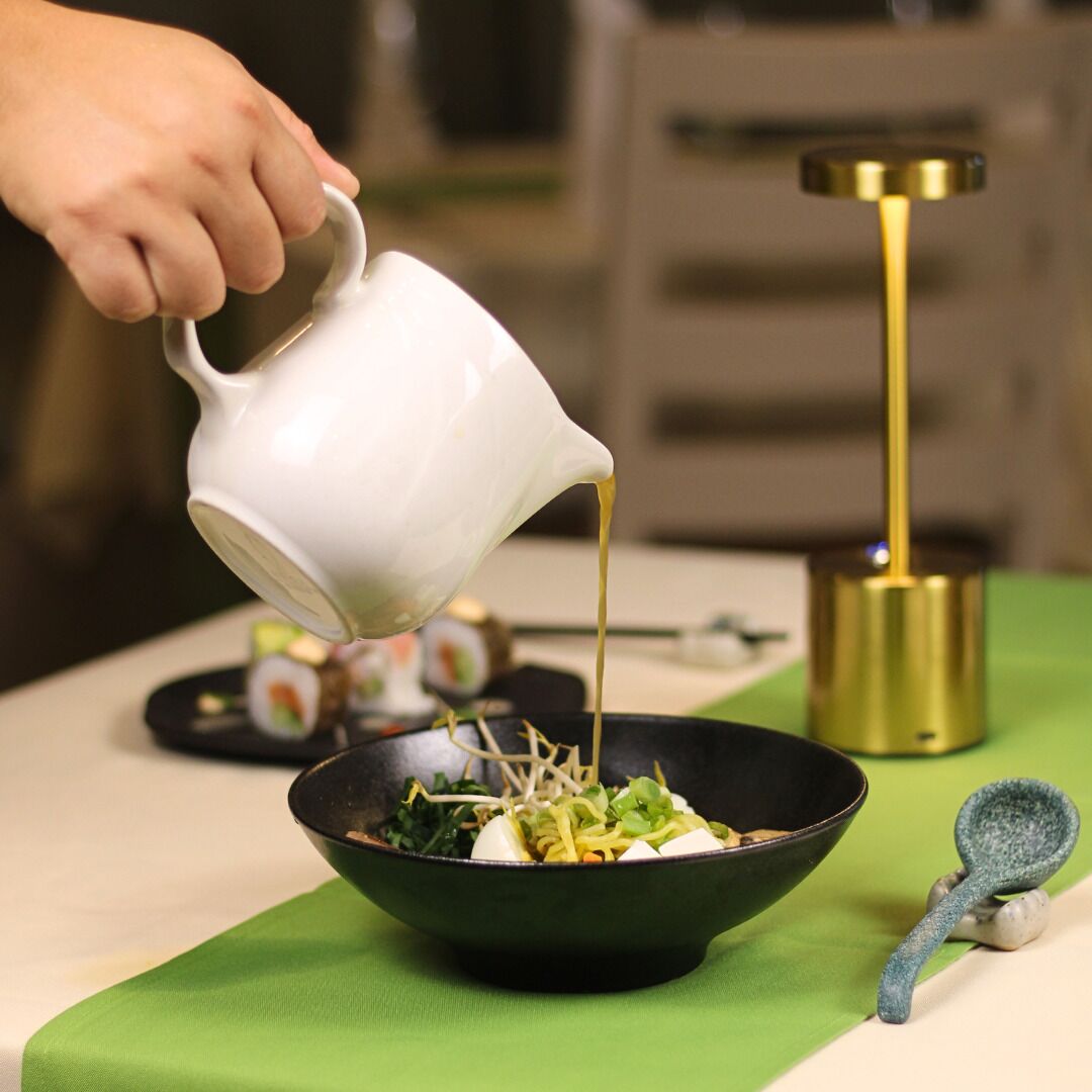 A person pouring a liquid from a white pitcher into a black bowl on a table with a green cloth.