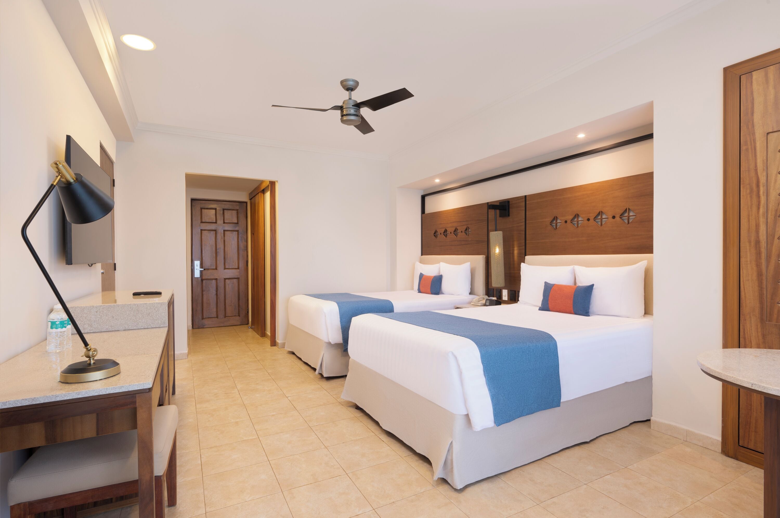 Une chambre d’hôtel avec deux lits, un bureau avec une lampe, un ventilateur de plafond et une tête de lit en bois agrémentée d’éléments décoratifs.
