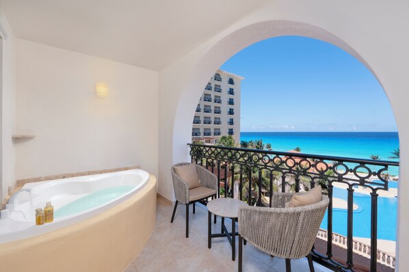 A balcony with a bathtub, two wicker chairs, and a view of the ocean and a hotel building.