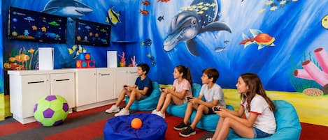 Ein Zimmer mit spielenden Kindern, die Videospiele spielen, einem großen Wandgemälde mit Meereslebewesen und einer weißen Kommode mit bunten Bauklötzen.