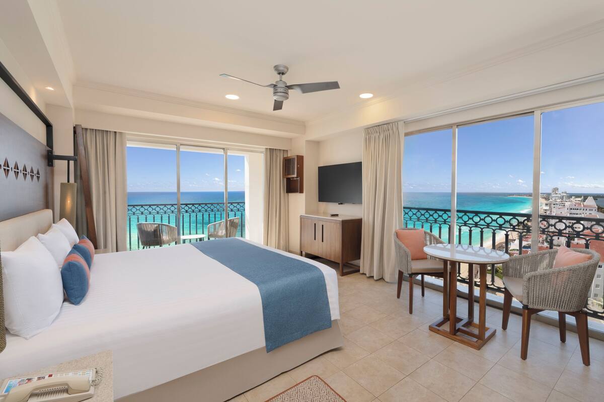 A hotel room with a bed, a desk, a chair, a TV, and a balcony with a view of the ocean.