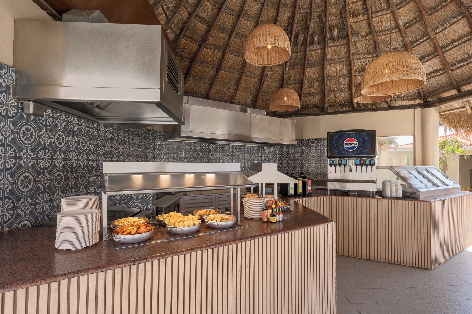A buffet setup with various food items, a Pepsi dispenser, and a thatched roof.